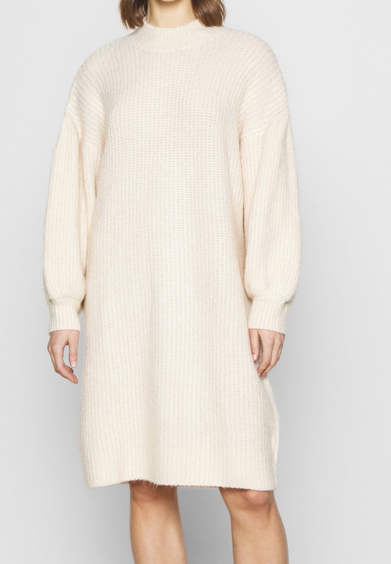 Femme portant une robe-pull en tricot ample, à manches longues, de couleur crème, avec des poignets et un ourlet côtelés, debout devant un fond uni.