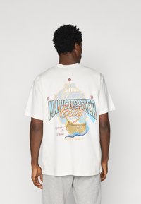 Camiseta blanca de algodón con un diseño gráfico en la parte posterior, que muestra "Manchester City" en azul y rojo con coordenadas geográficas.