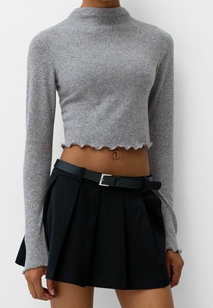 Pull court gris à manches longues avec bords volantés, associé à une jupe plissée noire taille haute et une ceinture noire.
