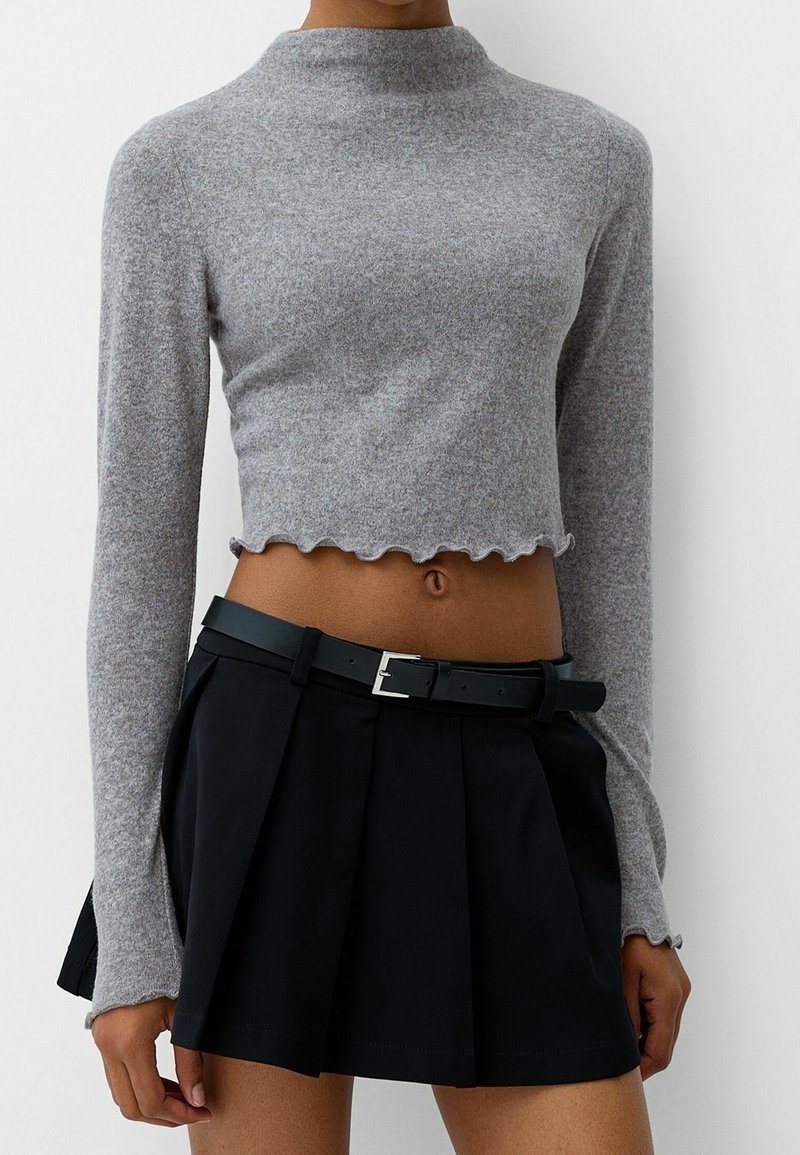 Pull court gris à manches longues avec bords volantés, associé à une jupe plissée noire taille haute et une ceinture noire.