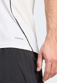 Hvid træningsskjorte med en sort søm og "AEROREADY" label ved kanten, parret med sorte textured shorts. Glat stof, tætsiddende design.