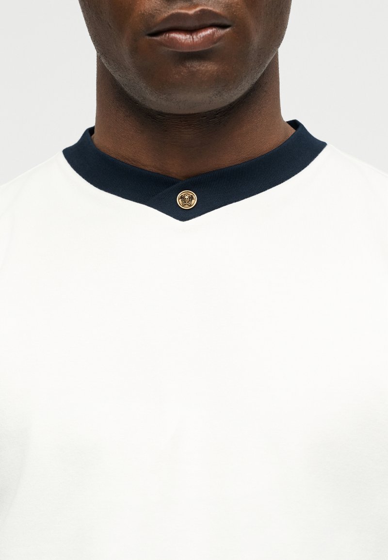 Close-up of a man wearing a white shirt with a navy collar fastened by a gold decorative button at the front center.