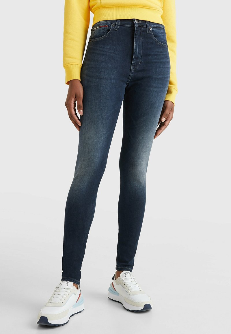 Jeans ajustados de tiro alto, de color azul oscuro con un lavado desgastado, que cuentan con cinco bolsillos y un cierre de botón frontal, combinados con zapatillas blancas.