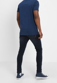 Marineblaues T-Shirt, eng anliegende dunkle Jeans und blaue High-Top-Sneaker. Merkmale sind die schlanke Passform und sichtbare Nähte an den Jeans.
