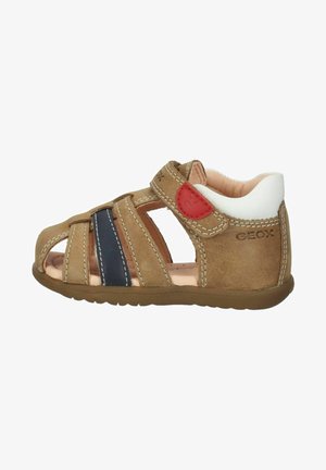 Sandal in pelle marrone con lati aperti, strisce blu navy, dettagli bianchi e cinturino in Velcro. Fodera interna morbida e suola in gomma. Design casual.