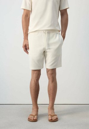 Hombre con pantalones cortos beige, camisa de manga corta a juego y sandalias marrones de pie frente a un fondo liso.