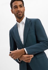Donkerturquoise maatpak met een notch lapel, gecombineerd met een witte overhemd. Bevat vier knopen bij de manchetten en een borstzak.