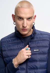 Jūros tamsiai mėlynas pūkuotas liemenė su vertikaliais siūlėmis, ant kurios yra "BIG STAR" logotipas, dėvima ant tamsiai mėlynos megztinio. Papuošalai - sidabro spalvos su turkio akmenimis.