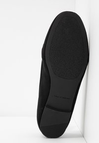 Semelle plate noire avec grip texturé et marque "CALL IT SPRING", positionnée à la verticale contre un mur d'angle blanc.