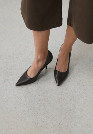 Femme portant des escarpins kitten à bout pointu en cuir noir texturé, associés à un pantalon marron large et raccourci, sur un sol gris clair.