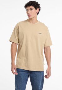 Beige kokvilnas T-kreklis ar īsām piedurknēm, apaļu kakla izgriezumu un melnu "GUESS JEANS" logotipu kreisajā krūšu daļā. Valkāts ar zilu džinsu biksēm.