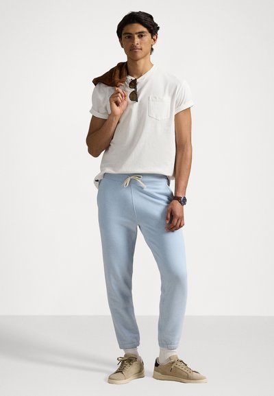 T-shirt en coton blanc avec poche, joggeurs bleu clair avec cordon de serrage, baskets beiges et veste marron portée sur l'épaule.