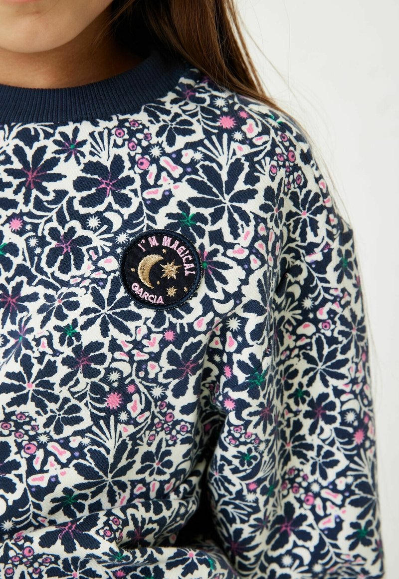 Gros plan sur un pull à motif floral en bleu marine, blanc et rose avec un patch rond portant l'inscription "I'M MAGICAL" orné de broderies de lune et d'étoiles.