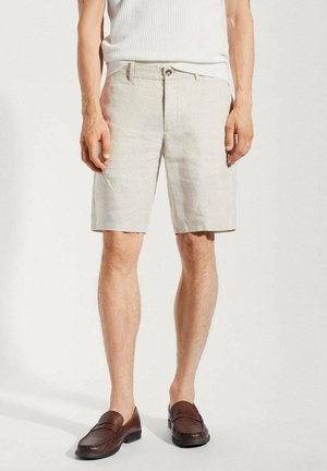 Hombre que lleva pantalones cortos beige hasta la rodilla, camiseta blanca sin mangas y mocasines de cuero marrón, de pie frente a un fondo claro.