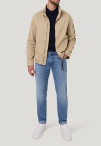 Veste légère beige avec un col haut, polo navy en dessous, jean bleu et baskets blanches. Look décontracté, peu de détails sur la veste.