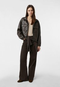 Brązowa kurtka ze sztucznej skóry z dwoma przednimi kieszeniami, noszona na beżowej bluzce i brązowych spodniach o szerokich nogawkach, z węzłowym detalem w talii.