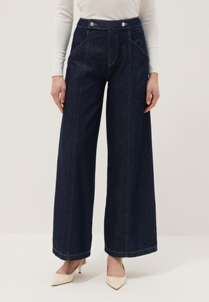Pantalones vaqueros de mezclilla azul oscuro de pierna ancha con cintura alta, bolsillos delanteros y detalles de costuras, combinados con tacones blancos de punta afilada y una blusa blanca.
