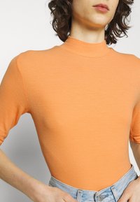 Personne portant un haut orange côtelé à manches courtes et ajusté, rentré dans un jean taille haute bleu clair, visible du cou à la taille.