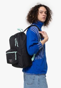 Zaino nero realizzato in tessuto resistente con cerniere e dettagli azzurri. Presenta una tasca anteriore e spallacci regolabili. Indossato sulla spalla.