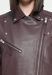 Veste en cuir bordeaux avec un col pointu, des éléments en métal argenté, une poche zippée diagonale et une fermeture éclair sur le devant. Texture lisse et coupe ajustée.