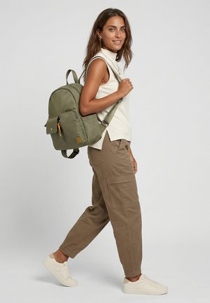 Kvinde går, bærer olivengrøn rygsæk, iført hvid ærmeløs top, brune cargobukser og hvide sneakers mod ensfarvet baggrund.