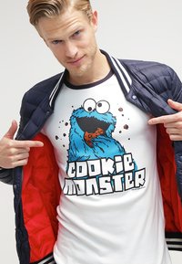 Weißes T-Shirt mit kurzen Ärmeln, das ein blaues, pelziges Cookie Monster-Design mit einem Keks und dem auffälligen Text "COOKIE MONSTER" zeigt, getragen unter einer marineblauen gesteppte Jacke.