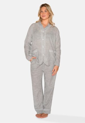 Vrouw staat op blote voeten en draagt een grijs-wit gestreept pyjamabovenstuk met knopen en bijpassende broek met zakken.