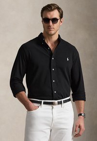 Hombre con camisa negra abotonada con logo, pantalones blancos, cinturón negro, reloj de pulsera y gafas de sol oscuras, de pie con una mano en el bolsillo.