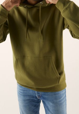 Person, die die Kapuze eines olivgrünen Hoodies anpasst, trägt hellblaue Jeans vor einem schlichten hellen Hintergrund.