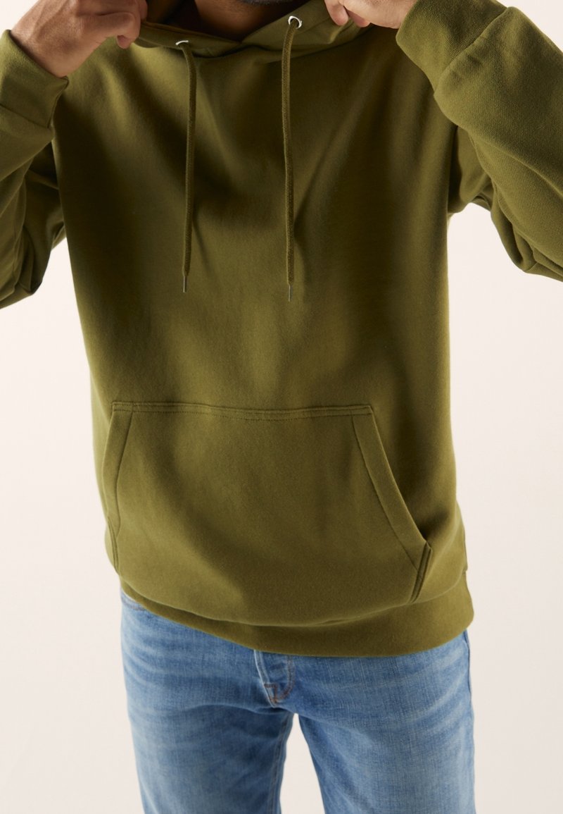 Personne ajustant la capuche d'un sweat à capuche vert olive, portant un jean bleu clair, sur un fond clair uni.