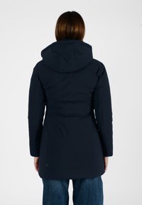 Femme portant un manteau d'hiver à capuche bleu marine et un jean bleu, se tenant de dos à l'appareil photo sur un fond blanc uni.