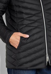 Hand met een lichte huidskleur in de linkerzak van een zwarte gewatteerde jas met een zilveren rits, waarbij de onderkant van een spijkerbroek zichtbaar is.