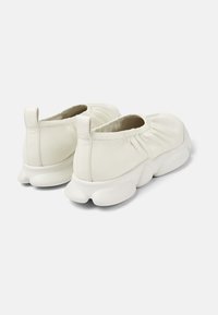 Scarpe bianche senza lacci con lati elasticizzati arricciati e suole spesse e testurizzate, viste da dietro su uno sfondo neutro.