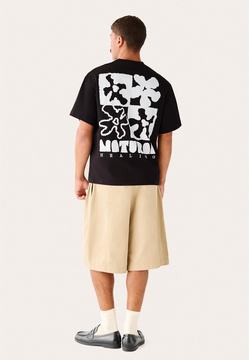 Homme portant un T-shirt graphique noir avec des formes abstraites blanches et le texte « Natural Healing », short beige, chaussettes blanches et mocassins noirs, vu de dos.