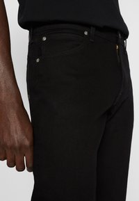 Gros plan sur une personne portant un jean en denim noir et une chemise noire, mettant en évidence la zone de la hanche et du haut de la cuisse avec une main visible.