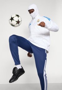 Witte hooded jack met blauwe strepen, marineblauwe sportbroek, zwarte voetbalschoenen en een voetbal die dicht bij het been is verhoogd.