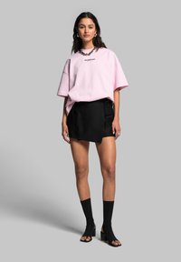 Svetle růžové oversized tričko s černým textem, zkombinované s černou sukní se zúženým detailem. Černé kotníkové ponožky a otevřené lodičky dokončují vzhled.