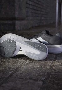 Par sorte og hvide løbesko på stenbelægning, den ene sko viser den teksturerede sål med "ADIZERO" branding.