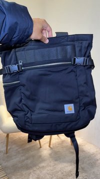 Zaino in tessuto nero con manico superiore, cerniere frontali e spallacci regolabili. Dotato di una toppa con logo e di un design strutturato.