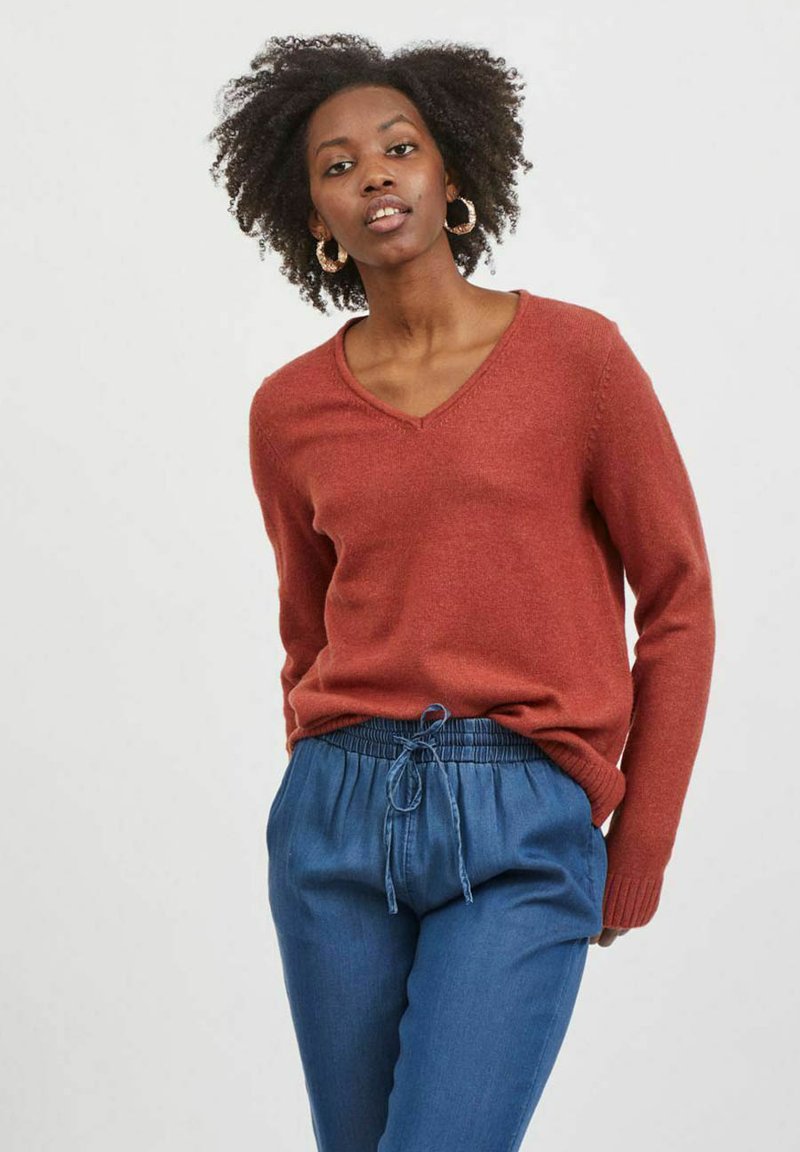 Pull en maille doux couleur rouille avec un col en V, associé à un pantalon bleu à cordon de serrage ample. Tissu texturé, design décontracté.