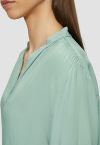 Hellgrüne Bluse mit weichem, glattem Textil, Mandarin-Kragen und Raffung entlang der Schulternaht.