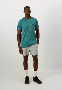 Mees, kes seisab valge tausta vastas, kandes teal värvi lühikeste varrukatega sportlikku särki, halli lühikesed pükse, valgeid sokke ja musti jooksujalatsit.