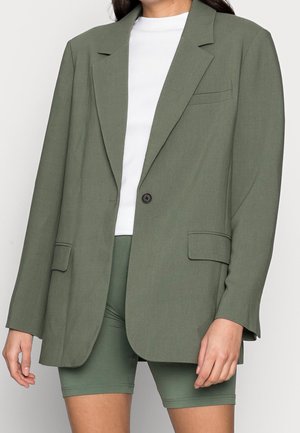 Vrouw draagt een oversized olijfgroene blazer over een witte top en bijpassende groene nauwsluitende korte broek, tegen een effen achtergrond.