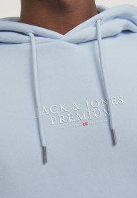 Svjetloplava dukserica s kapuljačom i vezicom, s utisnutim tekstom "JACK & JONES PREMIUM" u bijeloj boji. Izrađena od mekog materijala s ležernim krojem.