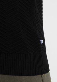 Zwarte gebreide trui met een chevronpatroon, geribde zoom en een klein logo-label. De stof lijkt zacht en gestructureerd.