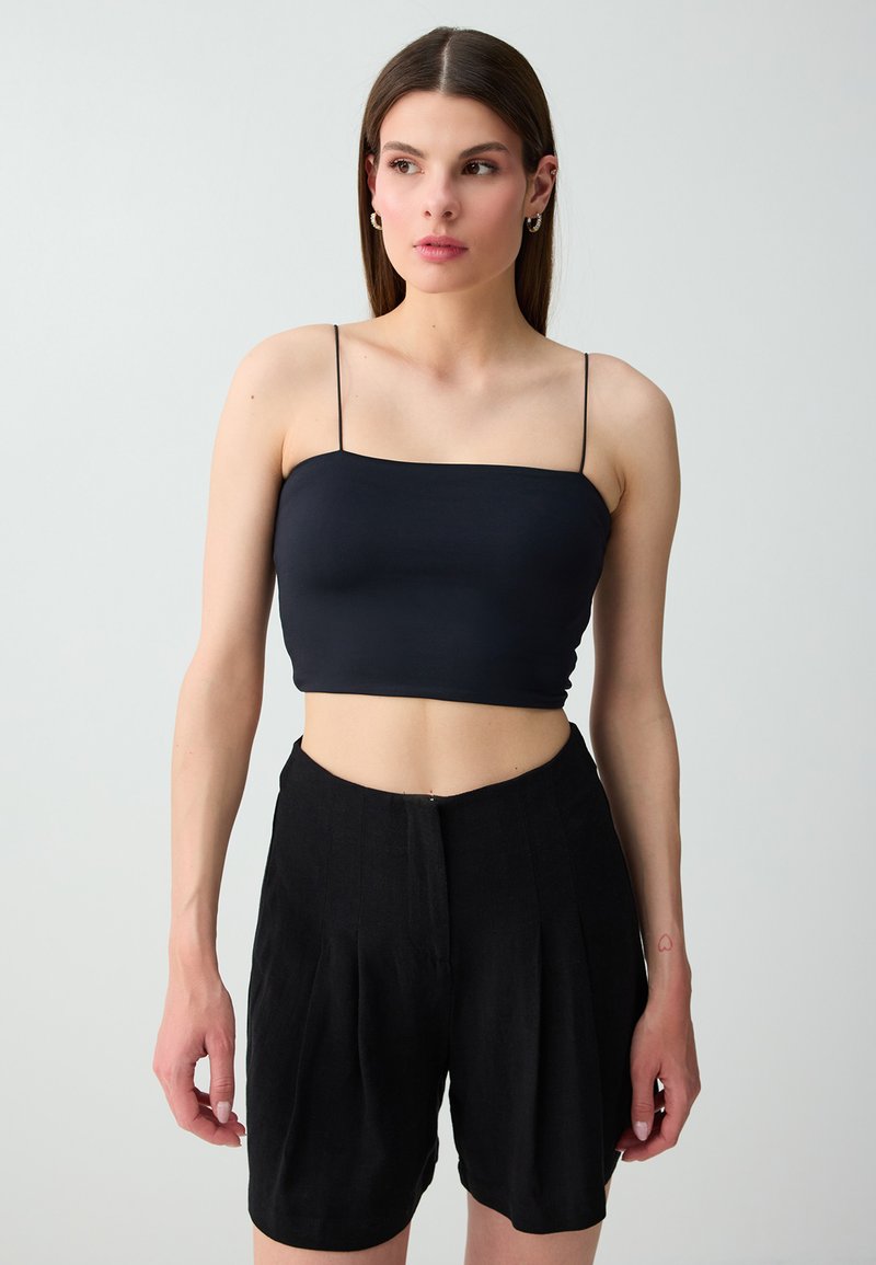 Jeune femme aux longs cheveux bruns portant un crop top noir à fines bretelles et un short noir taille haute sur un fond clair uni.