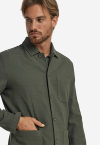 Grön texturerad jacka med krage, framknappar och bröstficka. Materialet har ett vävt utseende. Modellen har en hand i fickan.