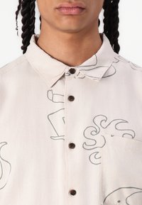 Camisetta a maniche corte con bottoni in tessuto beige chiaro con motivi astratti blu scuro, colletto e taschino sul petto con patta arrotondata.