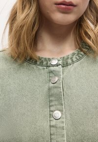 Ξανθό άτομο που φοράει ανοιχτοπράσινο πουκάμισο με ασημένια κουμπιά και στρογγυλή λαιμόκοψη, εμφανιζόμενο από το λαιμό έως το στήθος.