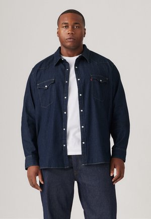 Donkerblauwe denim shirt met knoopsluitingen, twee borstzakken en een klassieke kraag. Draagt over een wit T-shirt met ronde hals.